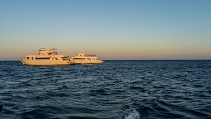 Obraz premium Two white yachts in the Red Sea are illuminated by the evening sun. Ripples on the blue water. Clear azure sky. The golden hour. Egypt
