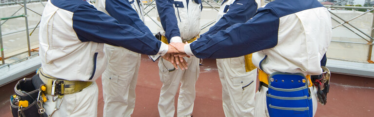 円陣を組んで手を重ね合わせる作業服姿の５人の男たち