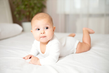 Portrait of a cute baby girl of 5 months in a white bodysuit on a white bed in the bedroom. Space for text. Child lifestyle