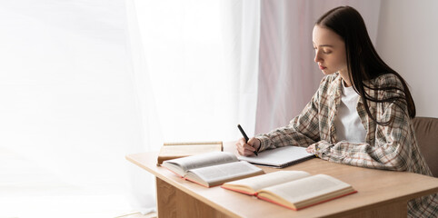 Purposeful female student studying with writing reference book, doing homework, concentrated teenage girl making notes in workbook, test or exam with textbooks, home school education concept