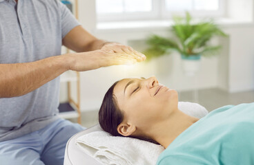 Happy relaxed and calm young woman with closed eyes receiving reiki treatment above head. Male therapist holding hands with yellow glowing light over female client's head lying on massage table.