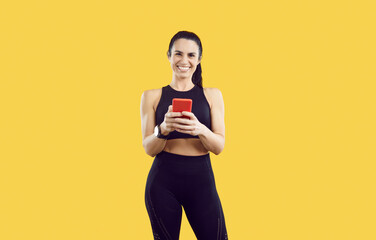 Portrait of joyful smiling fit woman with ponytail using smartwatch and fitness tracker on mobile phone. Happy beautiful lady in sports crop top checks workout app and is satisfied with her result