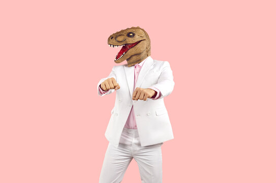 Weird guy in funny disguise dancing against pastel pink studio background. Cheerful eccentric man in white suit and silly ugly wacky masquerade dinosaur mask having fun at crazy party