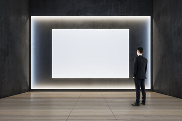 Businessman standing in modern exhibition hall interior with illuminated white mock up wall and wooden flooring. Gallery concept.