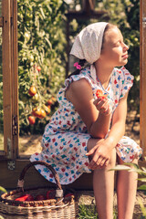 A girl in a summer dress and a kerchief is eating a tomato on the threshold of a greenhouse and...