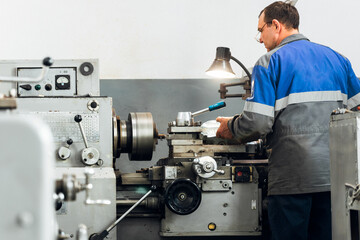 Turner stands behind lathe in production hall and works. View of worker from behind from back in...