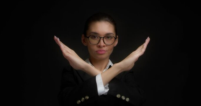 Angry woman in glasses and a business suit holds her arms crossed in a cross gesture and looks at the camera. The stop sign, the concept of negation, shows no on a black background