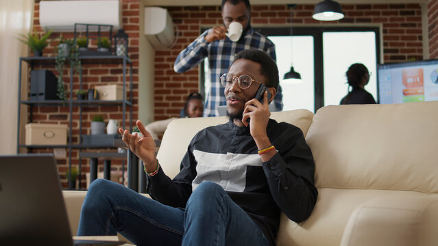 African american person chatting on phone call in business office, using smartphone to enjoy remote telecommunication with colleagues. Young man working on laptop and having conversation. - Powered by Adobe