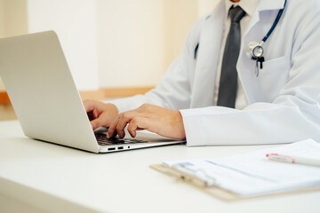 Closeup of male doctor expert wearing white coat using laptop and reading clipboard report at work in hospital.