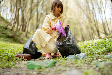 Fototapeta Young woman in protective clothes collecting scattered plastic garbage in the woods. Problem of bed ecology and environmental pollution with long-decomposing waste obraz