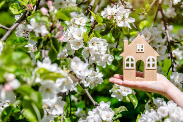 The girl holds the house symbol against the background of blossoming appletree