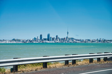 Auckland skyline on a sunny day, seen from a highway across the bay with parts of the street and the foreground and lots of blue turquoise water in between. 