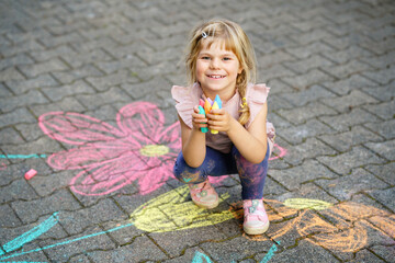 Little preschool girl painting with colorful chalks flowers on ground on backyard. Positive happy...