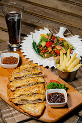 Meat casserole in puff pastry on a wooden stand with cups filled with sauces, against the background of a plate with green salad and a glass of dark juice.