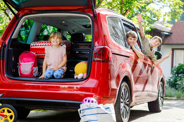 Three children, two boys and preschool girl sitting in car before leaving for summer vacations with parents. Happy kids, siblings, brothers and sister with suitcases and toys going on family road trip