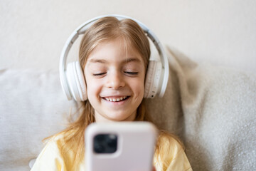 Cute child with headphones and mobile phone on sofa indoors.Smiling small child playing online game...