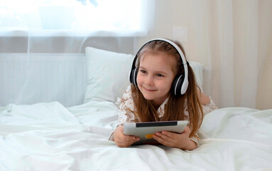 Concept of childhood, technology and people is a girl with a tablet computer and headphones lying on a bed at home. Online training. quarantine. The child listens to music with headphones and smiles