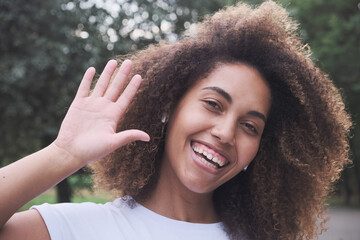 Closeup happy face biracial modern female in wireless earphones smiling waving hand outdoor at summer park. Portrait laughing mixed race curly woman natural beauty black skin greeting relaxing have
