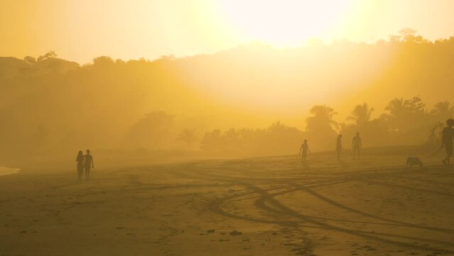 STILL SHOT: Silhouettes of unrecognizable people walking on misty beach at golden hour. Young couple and family enjoying strolling at Playa Venao beach. Leisure activities during summer holiday.