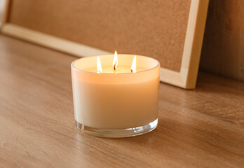 Obraz na płótnie Canvas A large scented candle with three burning wicks and melting wax in a glass, standing on a wooden table against the background of a board with cork notes. Side view. Place to copy