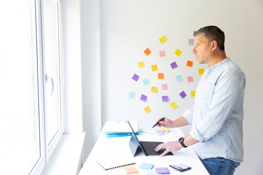 vMan with blue shirt stands at white height adjustable table and works on notebook behind him on the wall stick colorful notes