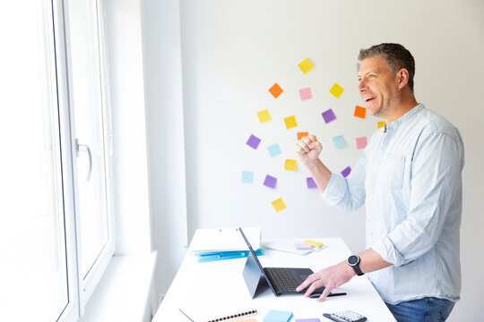 vMan with blue shirt stands at white height adjustable table and works on notebook behind him on the wall stick colorful notes
