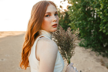 Side view of charming redhead woman with bouquet of grass standing on sand in nature in summer and looking at camera 