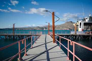 Entrance to jetty, on Lago General Carrera lake, Puerto Ibanez, Aysen, Chile. Pier with street lamps, mooring ropes crosses pier and moored ferry. Pier on glacial lake with mountain view in Patagonia