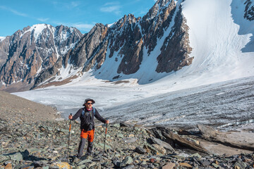 Hiker with trekking poles walks near large glacier tongue in high mountains. Scenic landscape with...