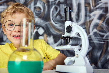 A transparent flask with a green liquid in the foreground covered half of the smiling boy's face. A satisfied child in round glasses and a bright T-shirt hides behind laboratory objects