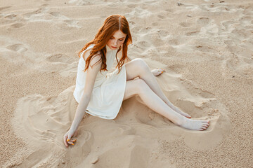 From above of barefoot woman with wavy red hair and in dress sitting on sandy beach and chilling in...