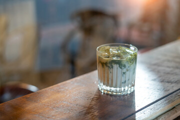A glass of Iced matcha latte green tea with ice and milk on a wooden bar over a cafe glass window reflex at a Cafe coffee shop. Cold brew refreshment summer drink with copy space. Selective focus