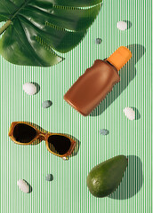 Tropical summer flat lay concept with stripped background, avocado, sunglasses, sunscreen, green leaf and stones.