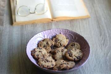 Bowl of chocolate chip cookies, open book and reading glasses on the table. Selective focus.