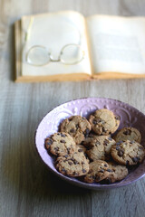 Bowl of chocolate chip cookies, open book and reading glasses on the table. Selective focus.