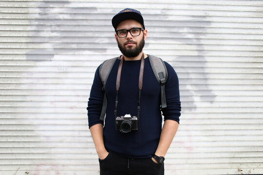 Street portrait of a male photographer with a beard in glasses and a cap with a vintage photo camera.