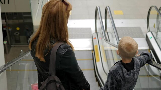 A mother and son with medical masks on their faces ride an escalator at the airport.