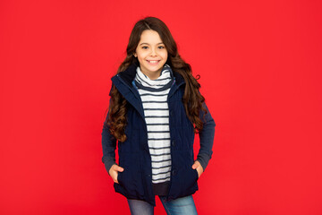 winter fashion. happy kid with curly hair in puffer waistcoat. teen girl on red background.