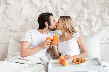 Loving young couple having breakfast in bed together, kissing, spending honeymoon at hotel