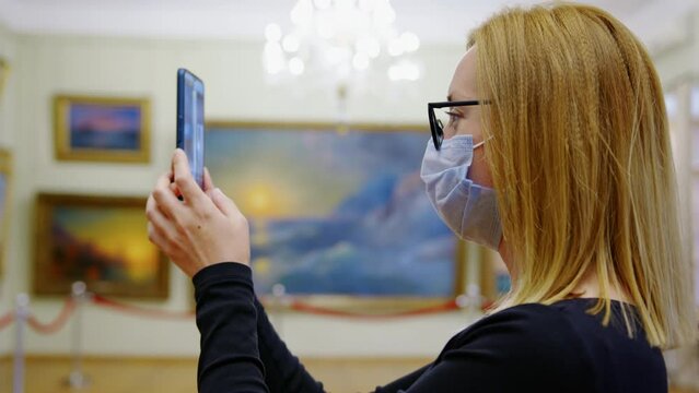 Blonde girl in mask and glasses takes pictures of paintings she likes in museum on smartphone. Young woman visits exhibition, cultural event keeping protection measures against coronavirus infection