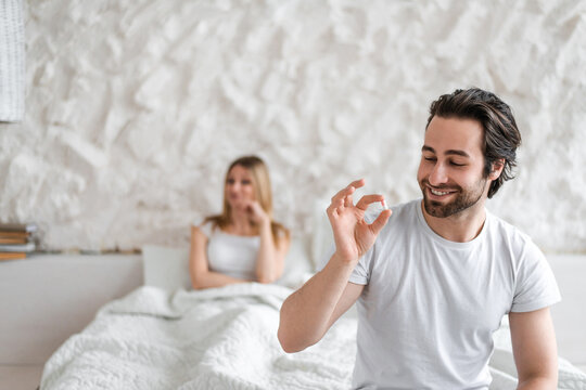 Young man holding pill, solving sexual dysfunction problems, happy woman sitting in bed and waiting for her husband