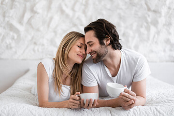 Romantic young couple lying on bed with cups of coffee, cuddling, spending time with each other at...
