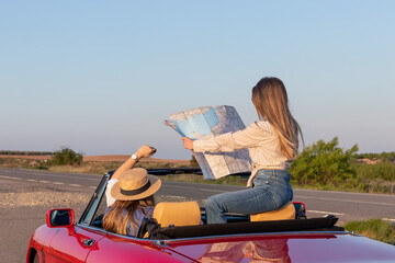 Women reading and holding a map sitting in red convertible car on remote road at sunset with golden...