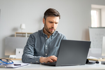 Mature male manager working on laptop at modern office, sitting at table, typing on computer keyboard
