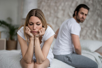 Stressed young woman sitting on bed, overwhelmed with family difficulties, her offended husband on...