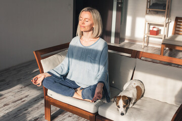 mindful middle aged woman making mudra gesture sitting in lotus position on couch at home