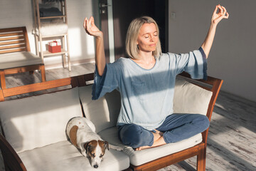 mindful middle aged woman making mudra gesture sitting in lotus position on couch at home