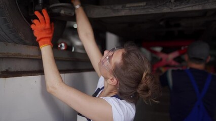 Female mechanic inspecting the car suspension. Strong woman coaching a male trainee. Worker checks car bottom and running gear of vehicle on lift. Workers in masks because of quarantine restrictions.