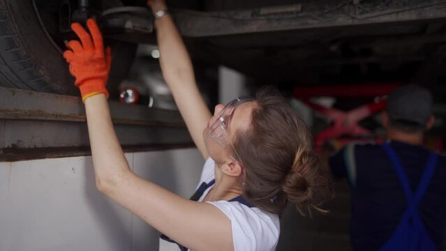 Female mechanic inspecting the car suspension. Strong woman coaching a male trainee. Worker checks car bottom and running gear of vehicle on lift. Workers in masks because of quarantine restrictions.