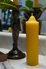 A tall emergency taper candle is displayed with a plant and candlestick in the background.